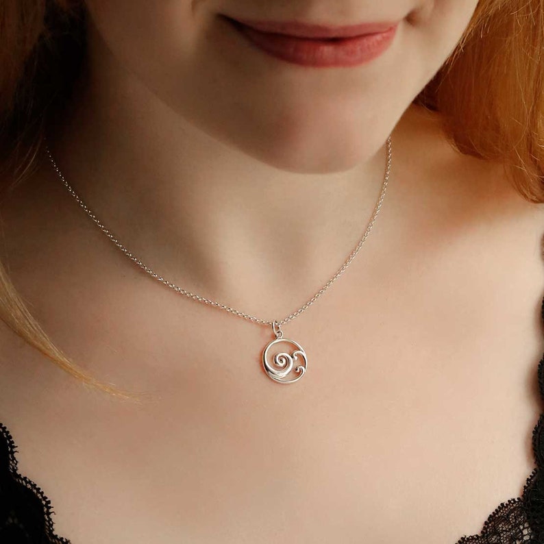 Puede incluir: Collar de plata con un colgante delicado. El colgante es un peque&ntilde;o c&iacute;rculo abierto con un dise&ntilde;o de olas en espiral en su interior. La cadena del collar es fina y plateada.