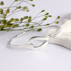 Może przedstawiać: Srebrna bransoletka typu bangle z zapięciem w kształcie łzy. Bransoletka wykonana jest z polerowanego metalu i ma młotkowaną fakturę. Zapięcie jest charakterystycznym elementem, dodającym biżuterii nowoczesnego charakteru. Tło obejmuje zieleń i białą powierzchnię.