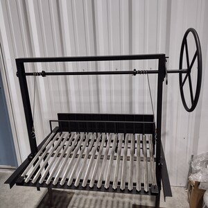 May include: A black metal grill with a stainless steel grate. The grill has a crank handle and is raised on two legs. The grill is in a warehouse setting.