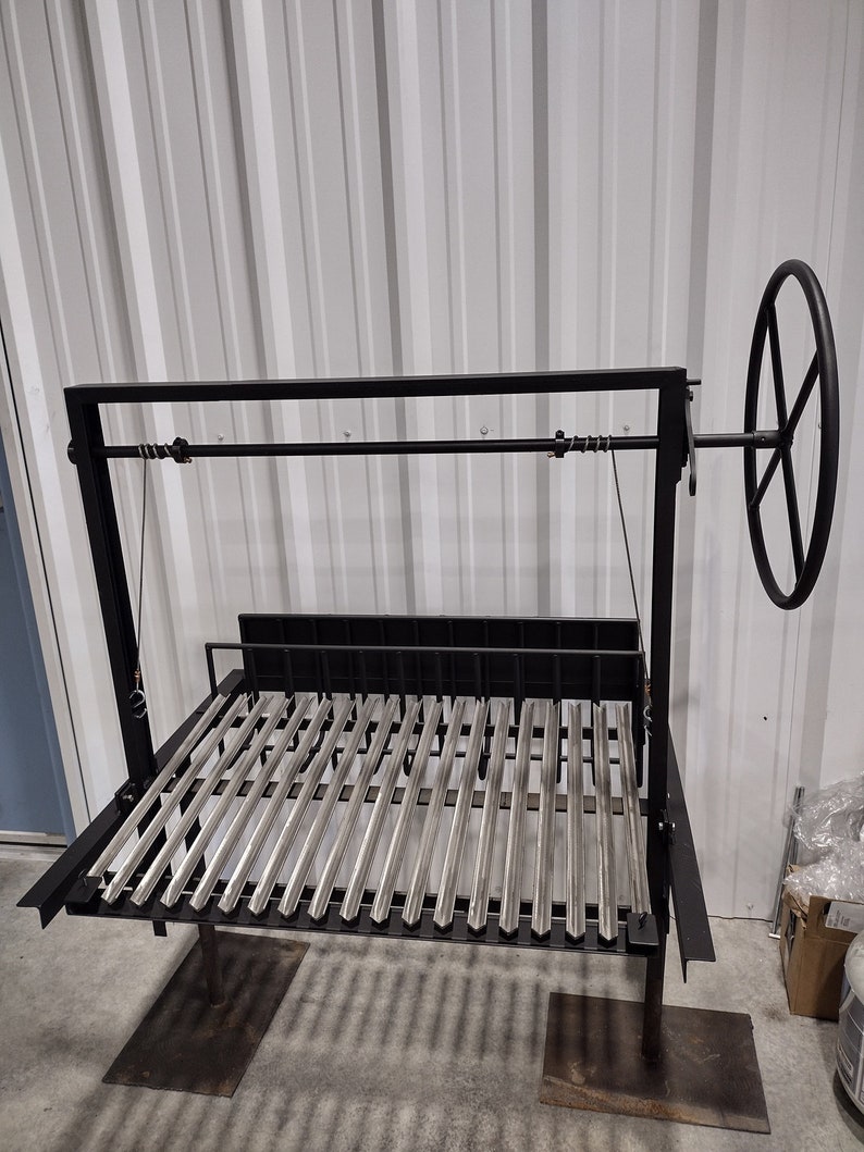 May include: A black metal grill with a stainless steel grate. The grill has a crank handle and is raised on two legs. The grill is in a warehouse setting.
