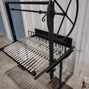 May include: A black metal grill with a silver grate and a large wheel for raising and lowering the grate. The grill is on a stand with a black base.