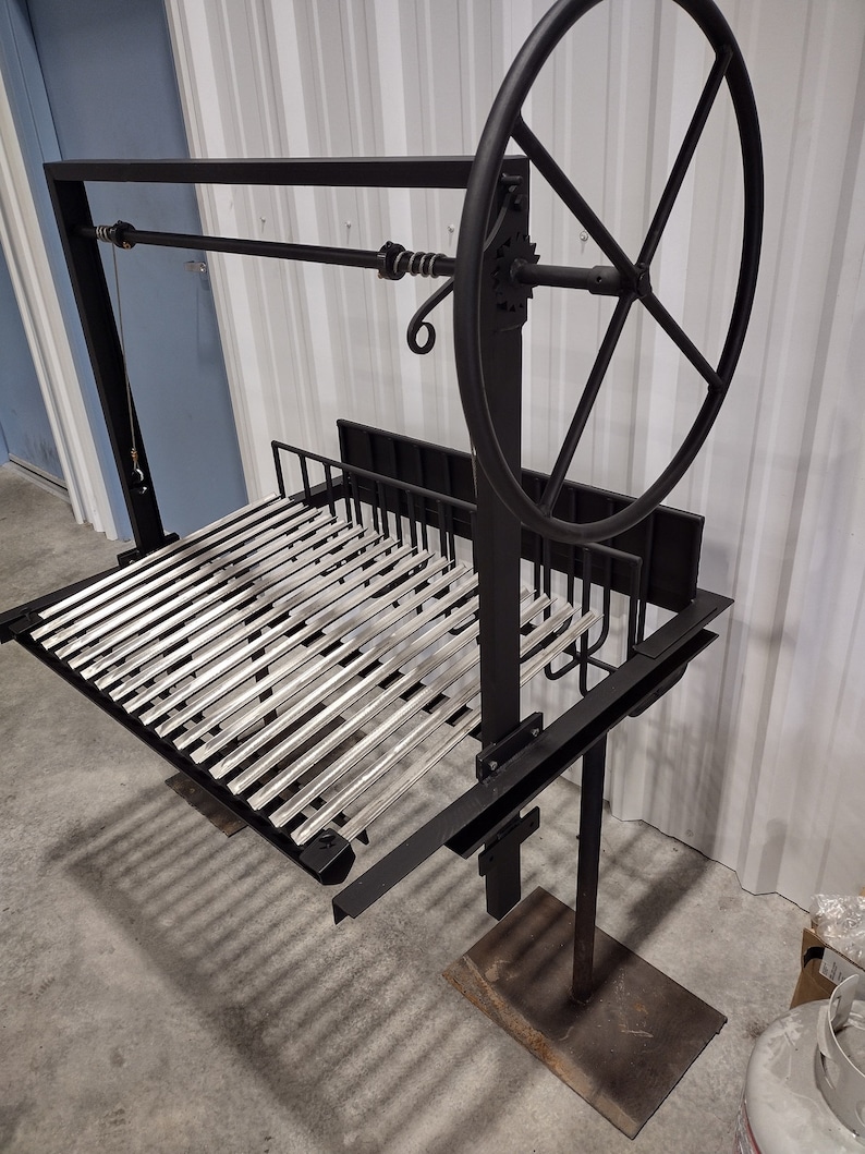May include: A black metal grill with a silver grate and a large wheel for raising and lowering the grate. The grill is on a stand with a black base.