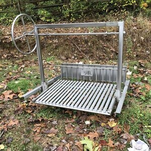 May include: A stainless steel outdoor grill with a rotating spit. The grill has a large, round wheel for turning the spit. The grill is set up on a grassy surface with brown leaves scattered around it.