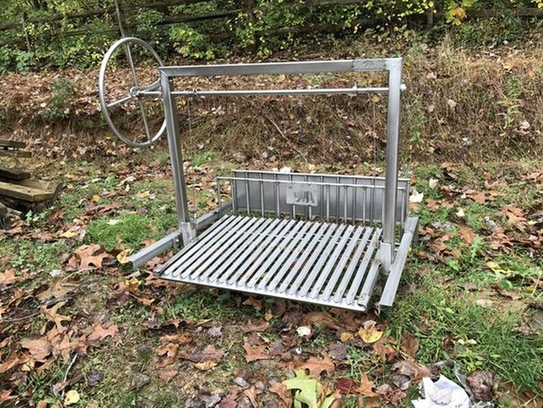 May include: A stainless steel outdoor grill with a rotating spit. The grill has a large, round wheel for turning the spit. The grill is set up on a grassy surface with brown leaves scattered around it.