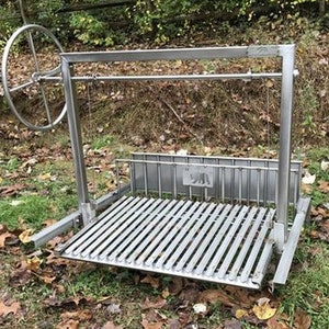 May include: A stainless steel outdoor grill with a rotating spit. The grill has a large rectangular cooking surface with a grate and a side shelf. The grill is on a grassy surface with fallen leaves.