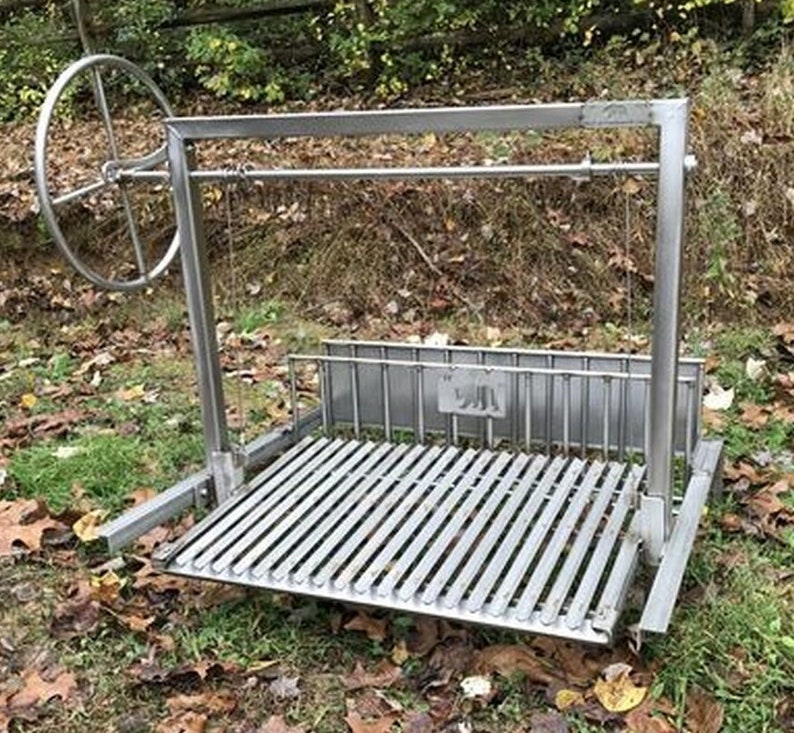 May include: A stainless steel outdoor grill with a rotating spit. The grill has a large rectangular cooking surface with a grate and a side shelf. The grill is on a grassy surface with fallen leaves.