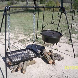 May include: A black metal tripod stand with a hanging chain and a cast iron pot. The stand also has a grill grate and a hook for hanging other items. The stand is set up over a pile of logs.