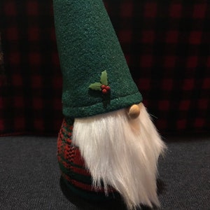 Peut inclure: Un gnome en feutre vert avec une barbe blanche et un corps en plaid rouge et vert. Le gnome porte un chapeau en feutre vert avec une feuille de houx et des baies rouges.