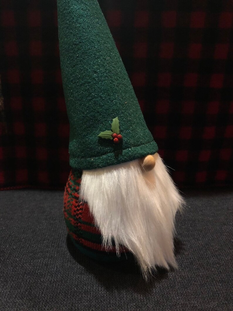 Peut inclure: Un gnome en feutre vert avec une barbe blanche et un corps en plaid rouge et vert. Le gnome porte un chapeau en feutre vert avec une feuille de houx et des baies rouges.