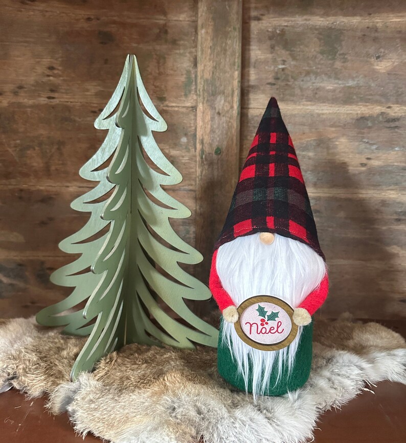 Christmas gnome with red, green, and black plaid hat, white beard, red arms, green body and holding a small Noel sign.