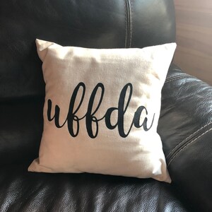 Könnte beinhalten: Ein weißes Kissen mit dem Wort "uffda" in schwarzer Kursivschrift.