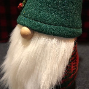 Peut inclure: Un chapeau de gnome en feutre vert avec une barbe en fourrure blanche et un corps en tartan rouge et vert.
