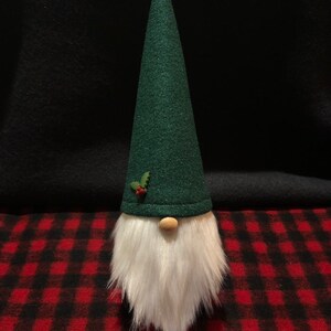Peut inclure: Un gnome en feutre vert avec une barbe blanche et une cloche dor&eacute;e sur le dessus de son chapeau. Le gnome est assis sur une surface &agrave; carreaux rouge et noir.