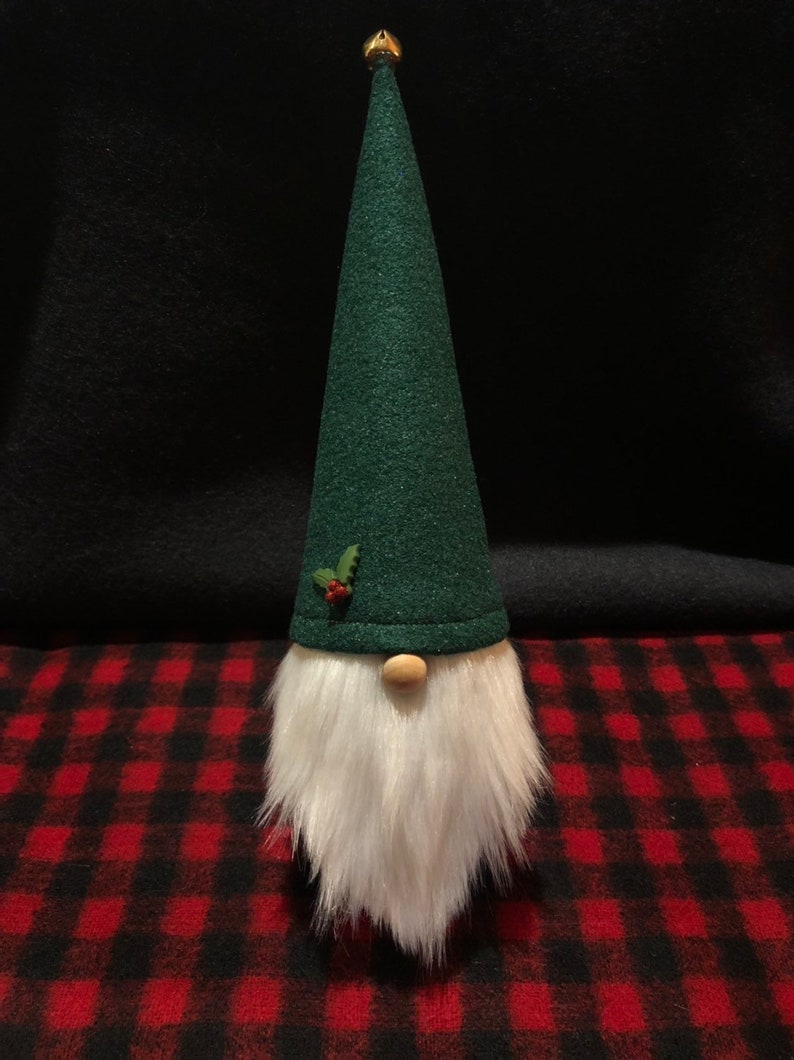 Peut inclure: Un gnome en feutre vert avec une barbe blanche et une cloche dor&eacute;e sur le dessus de son chapeau. Le gnome est assis sur une surface &agrave; carreaux rouge et noir.