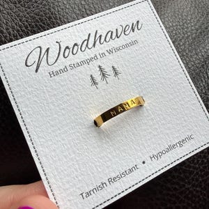 Pode incluir: Um anel dourado com a palavra "MAMA" estampada, apresentado num cartão branco. O cartão diz "Woodhaven Hand Stamped in Wisconsin" com um gráfico de árvore. O cartão também indica que o anel é resistente a manchas e hipoalergénico.