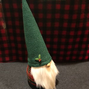 Peut inclure: Un gnome en tissu &agrave; carreaux verts et rouges avec un chapeau en feutre vert et une barbe blanche. Le chapeau est orn&eacute; d'une branche de houx avec des baies rouges.