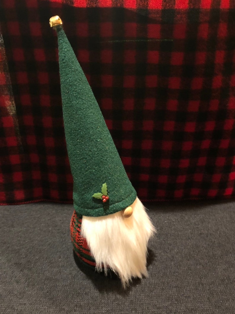 Peut inclure: Un gnome en tissu &agrave; carreaux verts et rouges avec un chapeau en feutre vert et une barbe blanche. Le chapeau est orn&eacute; d'une branche de houx avec des baies rouges.