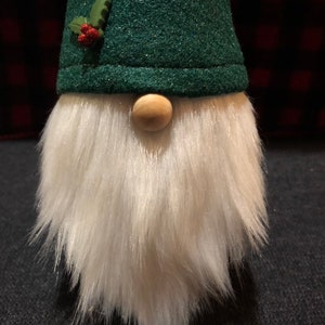 Peut inclure: Un chapeau de gnome en feutre vert avec une branche de houx et des baies rouges. Le gnome a une barbe blanche et moelleuse et un nez en bois.