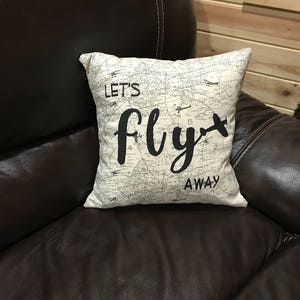 Może przedstawiać: Biała poduszka z wzorem przypominającym mapę i napisem "Let's fly away" czarnymi literami. Poduszka leży na brązowej sk&oacute;rzanej kanapie.