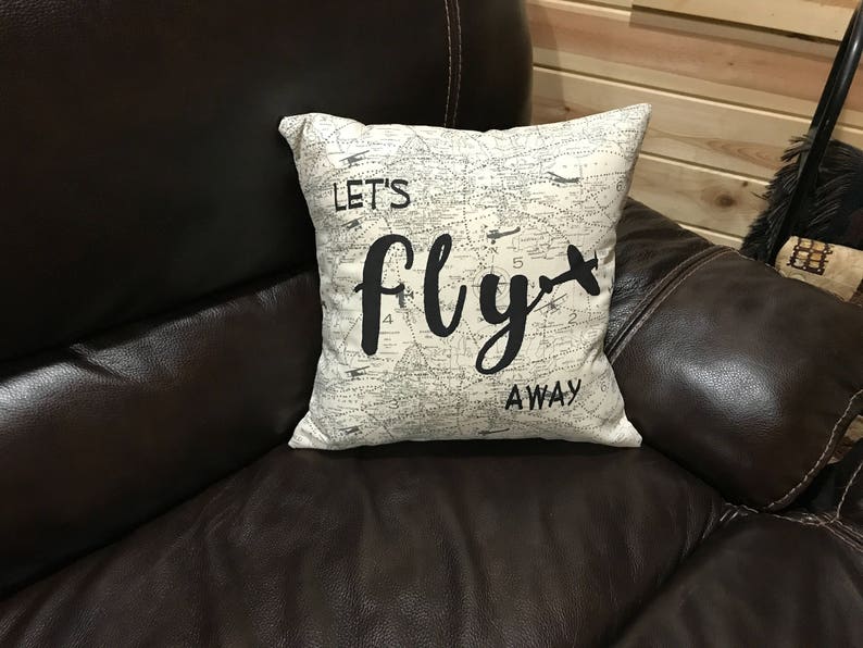Może przedstawiać: Biała poduszka z wzorem przypominającym mapę i napisem "Let's fly away" czarnymi literami. Poduszka leży na brązowej sk&oacute;rzanej kanapie.