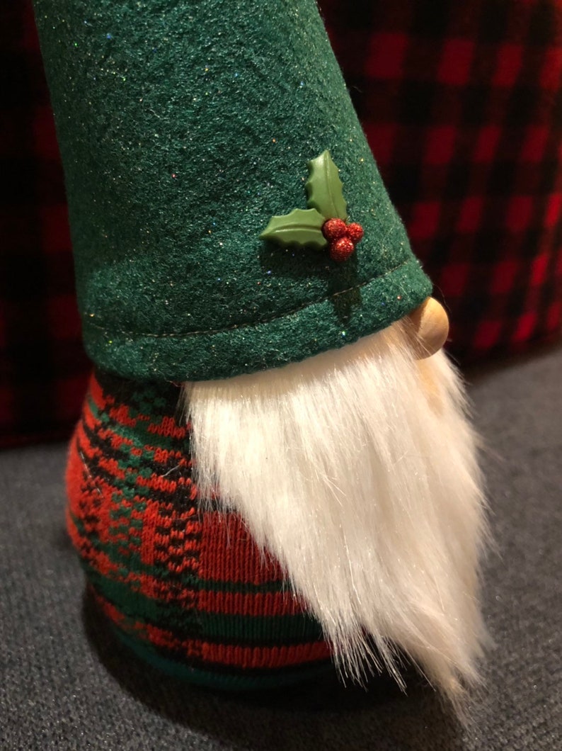Peut inclure: Un gnome en feutre vert avec une barbe blanche et un corps en tartan rouge et vert. Le gnome porte un chapeau en feutre vert avec une branche de houx et des baies rouges.
