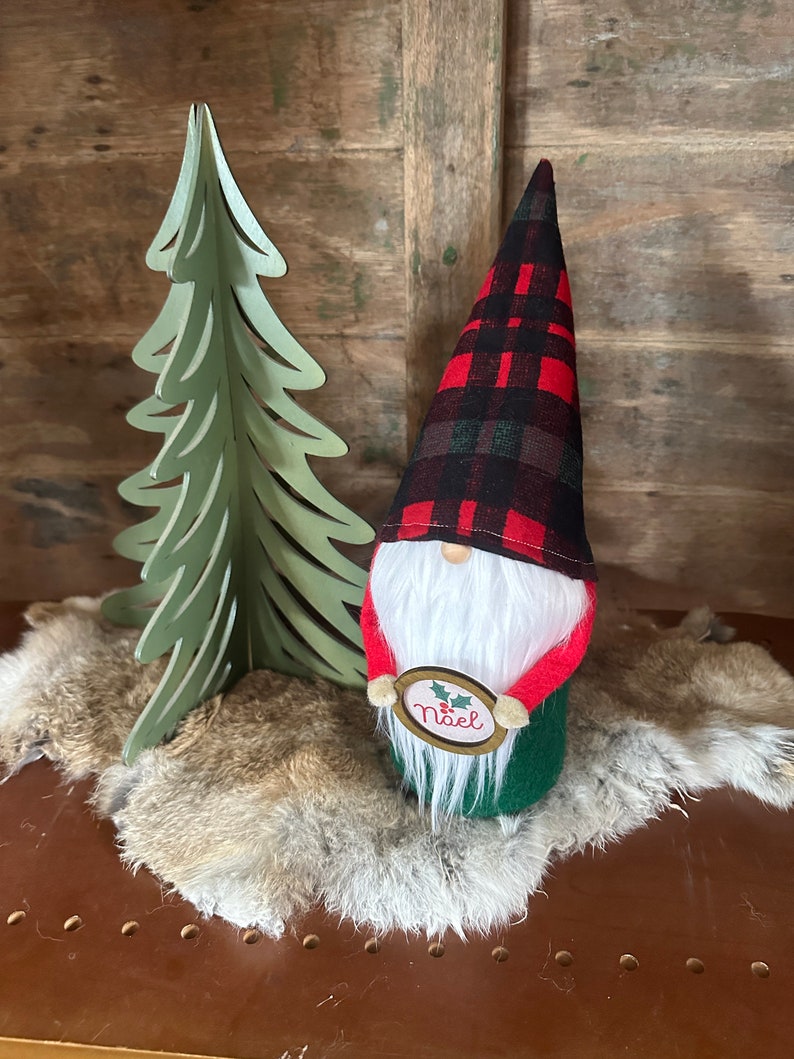 Christmas gnome with red, green, and black plaid hat, white beard, red arms, green body and holding a small Noel sign.