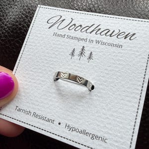 May include: A silver ring with two heart-shaped cutouts, presented on a white card. The card reads "Woodhaven Hand Stamped in Wisconsin" with a tree graphic. The card also states the ring is tarnish resistant and hypoallergenic.