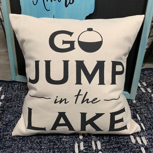 Puede incluir: Una almohada blanca con texto negro que dice "Go Jump in the Lake" y una silueta negra de un flotador de pesca.