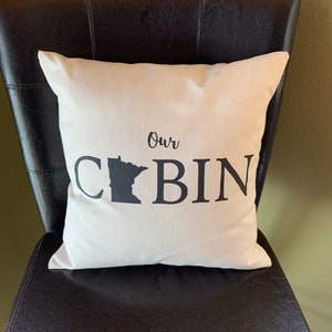 Puede incluir: Una almohada blanca con texto negro que dice "Our Cabin" y una silueta negra del estado de Minnesota. La almohada está sentada en una silla de cuero negro.