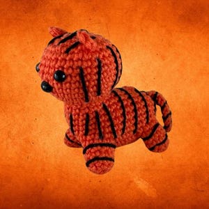 May include: A crocheted tiger figurine in orange with black stripes. The handmade toy has small black eyes and a tiny tail. The tiger is posed on an orange background.