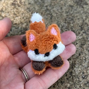 May include: A small, plush fox figurine featuring orange fur, a white chest and tail tip, and dark brown paws. The fox has embroidered pink inner ears and black eyes. The design is compact with a soft, fuzzy texture and rounded features.