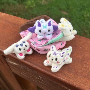 May include: A small pink fabric pouch with a brown top and a light green zipper, containing three white plush animal figures. Each plush features embroidered details in purple, blue, yellow, and pink, with black eyes and small ears.