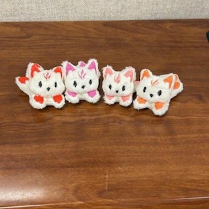 May include: Four small, white plush cat figures with colourful accents. Each cat has a different colour scheme, including red, pink, and orange. The cats are arranged in a row on a wooden surface, showcasing their detailed features and playful design.