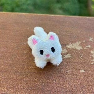 May include: A small, white, plush cat figurine with a fuzzy texture. The cat has embroidered black eyes, a small pink nose, and pink inner ears. It is depicted in a lying position with its tail curled slightly to the side.