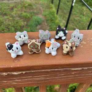 May include: A collection of eight small, plush cat figurines featuring various color patterns including white with black spots, grey, brown, and calico. Each miniature stuffed animal has embroidered facial features, small ears, and a soft, fuzzy texture.