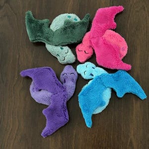 May include: A collection of plush dragon toys in various colors, including green, pink, purple, and blue. Each dragon has a soft, textured appearance with wings and a smiling face. The toys are arranged on a dark wooden surface.