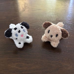 May include: Two small plush animal figures. One is white with black spots, black ears, and blue and pink embroidered details. The other is light brown with dark brown ears and black embroidered eyes and nose. Both have fuzzy textures.