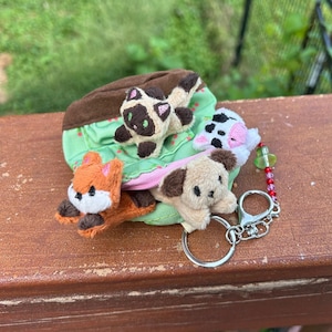May include: A small green fabric pouch featuring a brown trim and a pink zipper, adorned with four plush animal figures: a Siamese cat, a cow, a fox, and a dog. A silver metal keychain with a beaded charm is attached to the side of the pouch.
