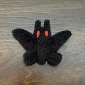Puede incluir: Una pequeña figura de peluche negra y difusa con forma de polilla o murciélago, con dos orejas prominentes y erguidas y dos ojos ovalados de color rojo brillante. La textura del material es suave y densa, cubriendo todo el cuerpo y las alas.