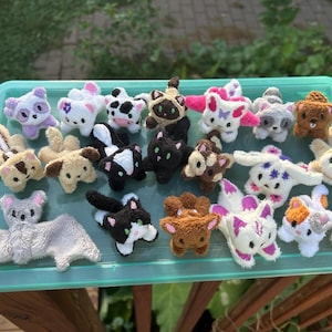 May include: A collection of small, handmade plush animal figures arranged on a teal plastic tray. The assortment includes various creatures such as cats, dogs, a bat, a skunk, and a raccoon, all crafted from soft, textured fabric in diverse colors.