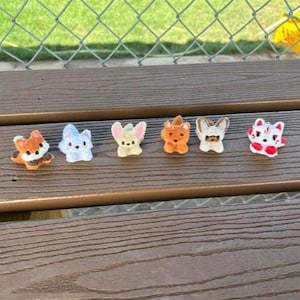 May include: A set of six small, plush animal figurines arranged in a row. The figures include an orange fox, a light blue creature, a tan fennec fox with large ears, a brown bear, a white and brown cat, and a white fox with red accents.