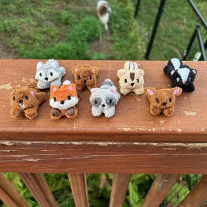 May include: A collection of eight small, plush animal figurines including a brown beaver, a grey wolf, a brown bear, a tan chipmunk, a black and white skunk, an orange fox, a grey raccoon, and a brown deer, all arranged on a wooden surface.
