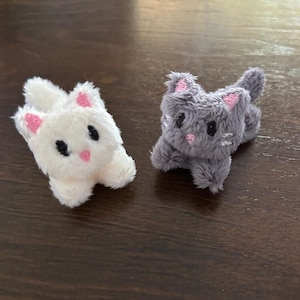 May include: Two small plush cat figurines, one white and one gray, featuring soft fuzzy textures, embroidered black eyes, pink stitched noses, and pink inner ears. Each miniature cat has a rounded body, short limbs, and a small tail, crafted with simple, clean lines and a compact design.