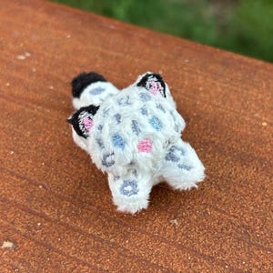 May include: A small, white embroidered patch shaped like a snow leopard cub. The design features a white base with grey, blue, and pink spots, black and pink ears, and a black-tipped tail, rendered in detailed thread work on a white fabric.