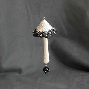 May include: A white and black mushroom ornament with a black bead hanging from the bottom. The mushroom is hanging from a string.