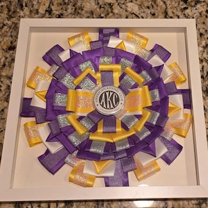 May include: A framed rosette featuring a circular design of purple, yellow, white, and silver ribbons. The center displays a circular emblem with the text "AMERICAN KENNEL CLUB" and "AKC FOUNDED 1884". The rosette is encased in a white frame.