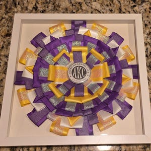 May include: A framed rosette featuring a circular design of layered ribbons in purple, yellow, white, and silver. The center displays a circular emblem with the text "AKC" and "American Kennel Club". The rosette is framed in a white square.