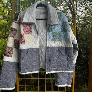May include: A quilted jacket featuring a patchwork design of various floral and paisley patterns in shades of blue, red, and white. The garment has a collared neckline, long sleeves, two front pockets, and a white interior lining.