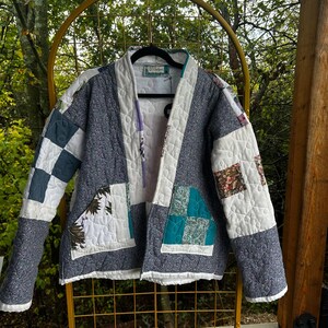 May include: A quilted jacket featuring a patchwork design with various fabric patterns including blue paisley, white, and teal squares. The garment has a shawl collar, long sleeves, and two front pockets, with a visible label reading Clothed.