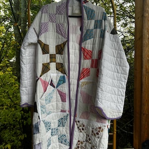 May include: A long-sleeved quilted coat made from white fabric with a patchwork design featuring various patterns and colors including plaid, floral, and gingham squares. The coat has a purple trim along the edges and two front pockets.